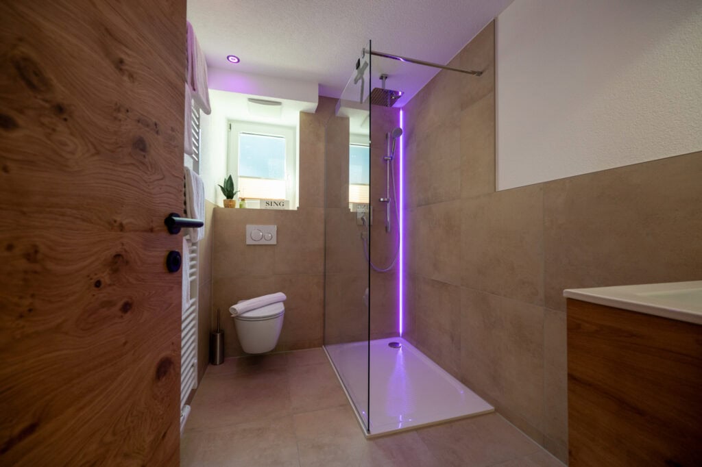 Modernes Badezimmer der Ferienwohnung Murmale mit bodengleicher Walk-in-Regendusche, stimmungsvoller violetter LED-Beleuchtung, beigen Fliesen und Holztür.