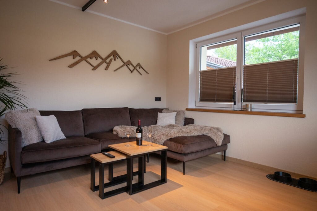 Gemütliche Couchecke in der Ferienwohnung Murmale mit braunem Ecksofa, Deko-Bergkette aus Holz, Couchtisch mit Wein und Hundeschüsseln auf hellem Holzboden.