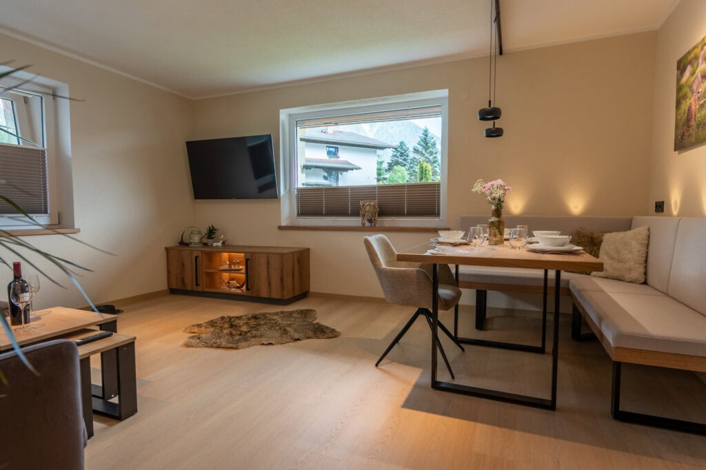 Offener Wohn- und Essbereich der Ferienwohnung Murmale mit moderner Eckbank, gedecktem Holztisch, Wand-TV und beleuchtetem Sideboard.