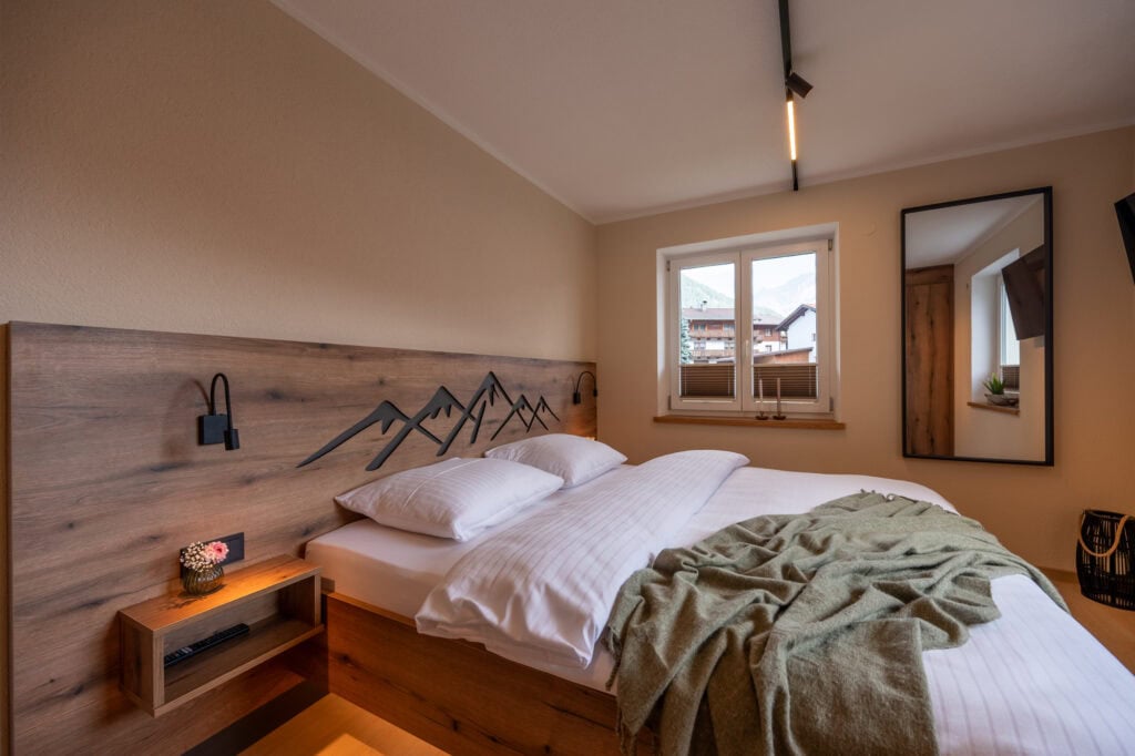 Gemütliches Schlafzimmer der Ferienwohnung Murmale mit Boxspringbett, Holz-Kopfteil mit Berggravur, Nachttischlampen und großem Spiegel.