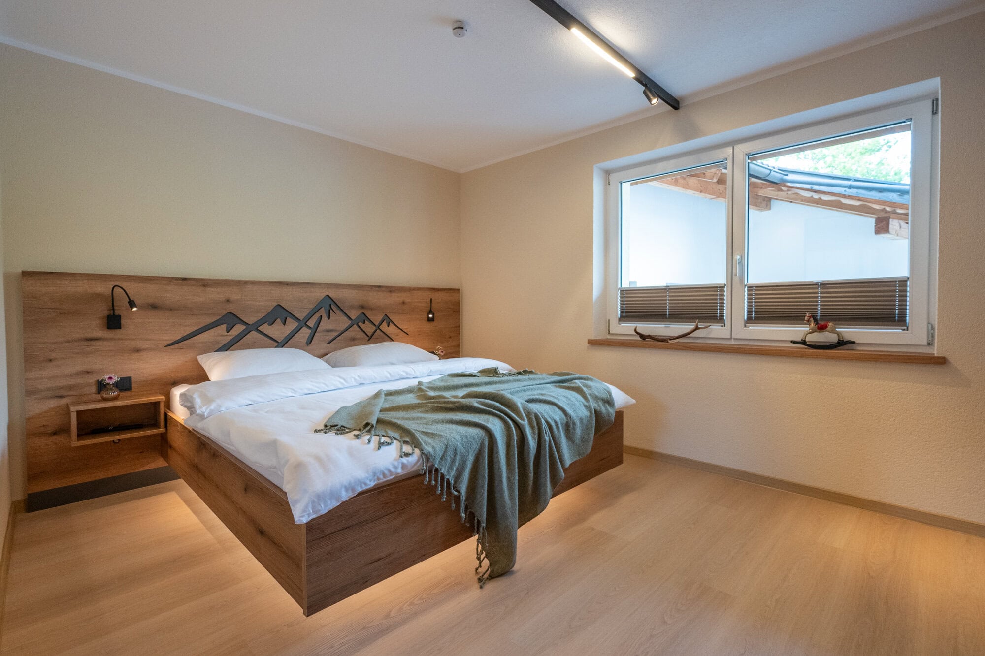 Schlafzimmer Vorne der Ferienwohnung Murmale mit Echtholzbett, Berg-Silhouette am Kopfteil, grünem Plaid und großem, schwarzen Wandspiegel.